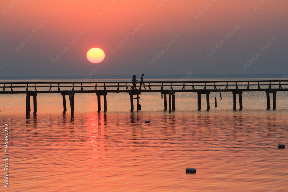 A couple walking down a long pier with the dropping sun in the background.
