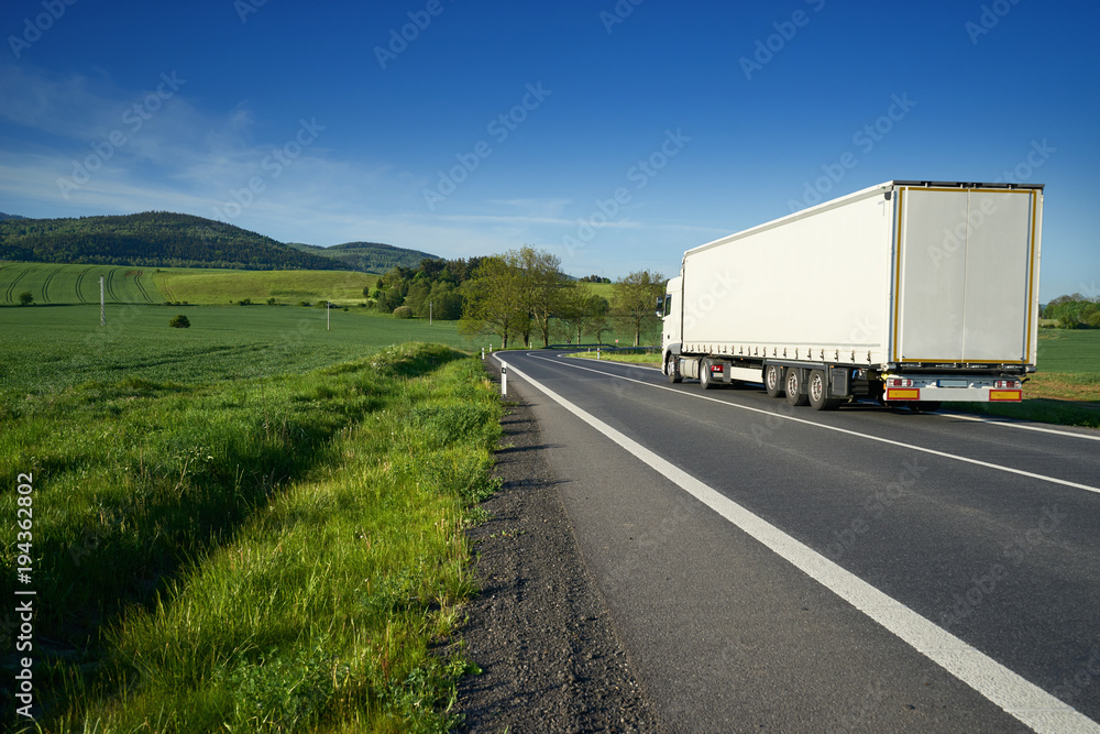 White truck on the asphalt road between green fields in rural landscape with wooded mountains in background