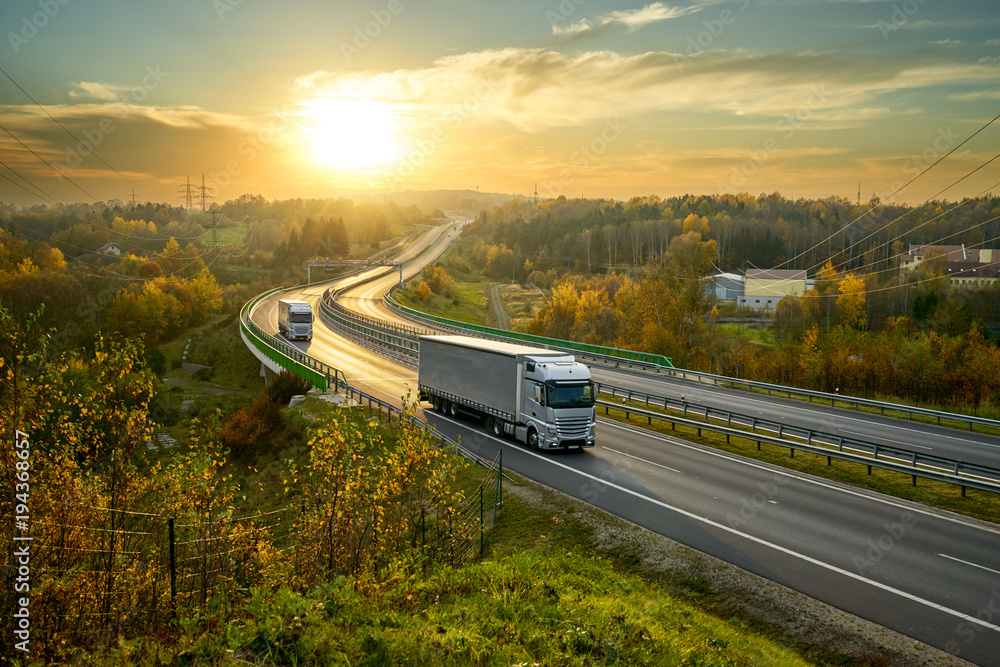 Silver trucks driving on the highway winding through forested landscape in autumn colors at sunset