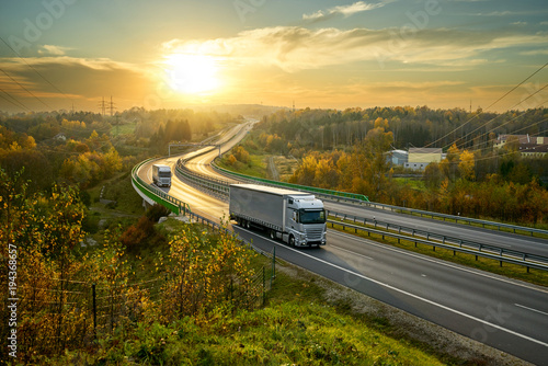 Obraz na płótnie Silver trucks driving on the highway winding through forested landscape in autumn colors at sunset w salonie