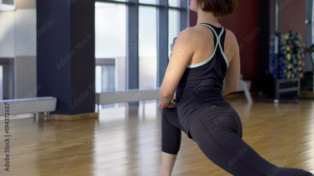 Woman in gym makes an exercise of lunging with legs for stretching. During Pilates classes, an altetic female person with a view of back is doing a healing workout on mat. Woman in gym makes an exercise of lunging with legs for stretching. During Pilates classes, an altetic female person with a view of back is doing a healing workout on mat.
