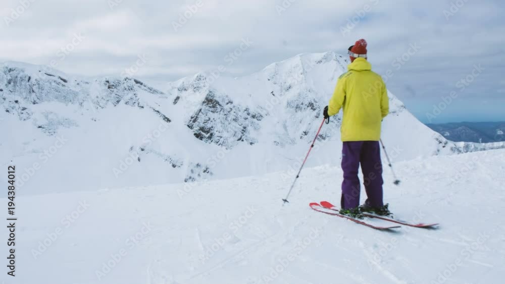 A skier in a bright suit comes to the top of the mountain, looks at the snowy mountains, 4k. A skier in a bright suit comes to the top of the mountain, looks at the snowy mountains, 4k.