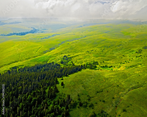 Aerial view of green hills. Summer landscape. Green grassy meadow on a hillside on top of mountain ridge with some forest under cloudy blue sky. Adygea  Lago-naki  Lagonaki  Krasnodar  Russia.