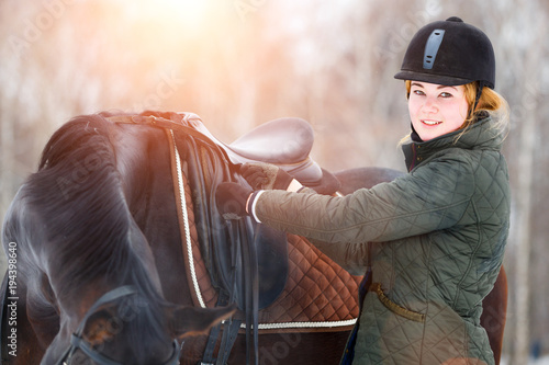 Young woman adjusting stirrups and saddle before riding her bay horse.