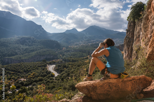 Man sits at the edge of a rock and looks at valley and mountains 
