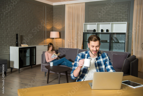 Man with cup in hand using laptop while woman reading book on couch in living room in modern design