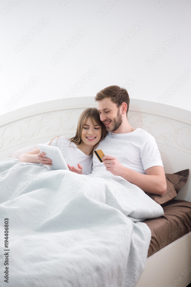 Fototapeta premium Smiling woman using digital tablet with her boyfriend holding credit card in cozy bedroom