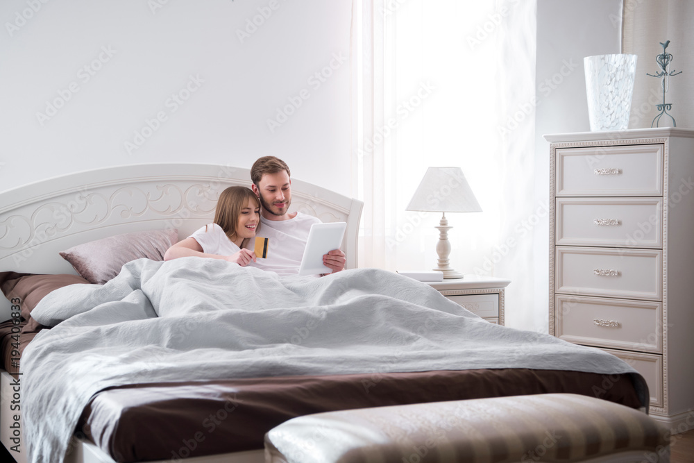 Fototapeta premium Young couple lying in bed and doing online shopping in modern bedroom