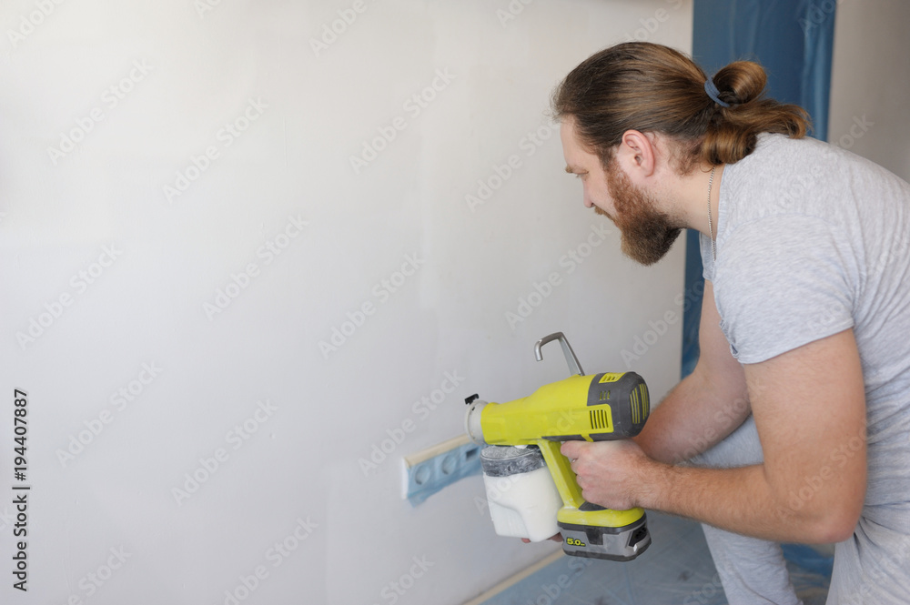 The man with a beard paints white walls with the paint spray. Repair in the house The man with a beard paints white walls with the paint spray. Repair in the house