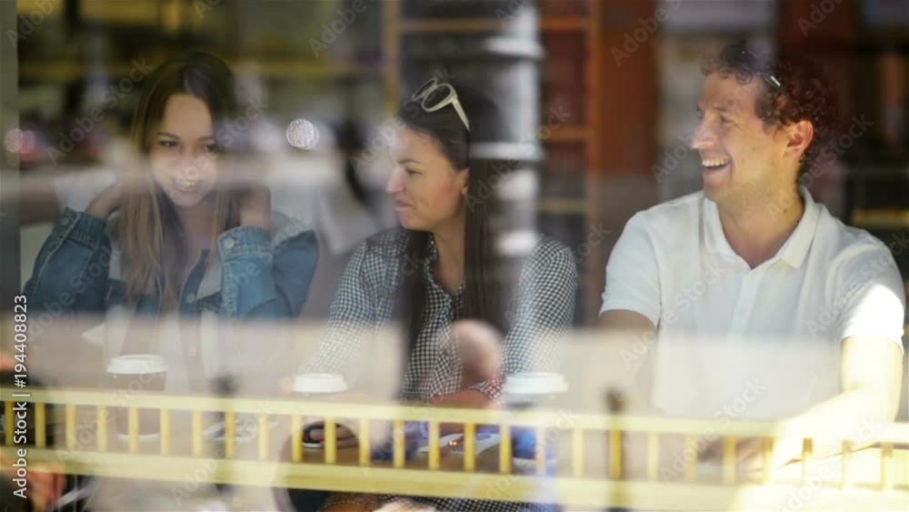 Group of Young People Having a Good Time at a Cafe and Waiting for Some Beverages. Two Women and One Man in Restaurant Seen Through Window on the Street. Group of Young People Having a Good Time at a Cafe and Waiting for Some Beverages. Two Women and One Man in Restaurant Seen Through Window on the Street.