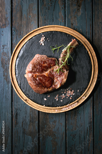 Grilled black angus beef tomahawk steak on bone served with salt  pepper and rosemary on round wooden slate cutting board over dark wooden plank background. Top view  copy space.