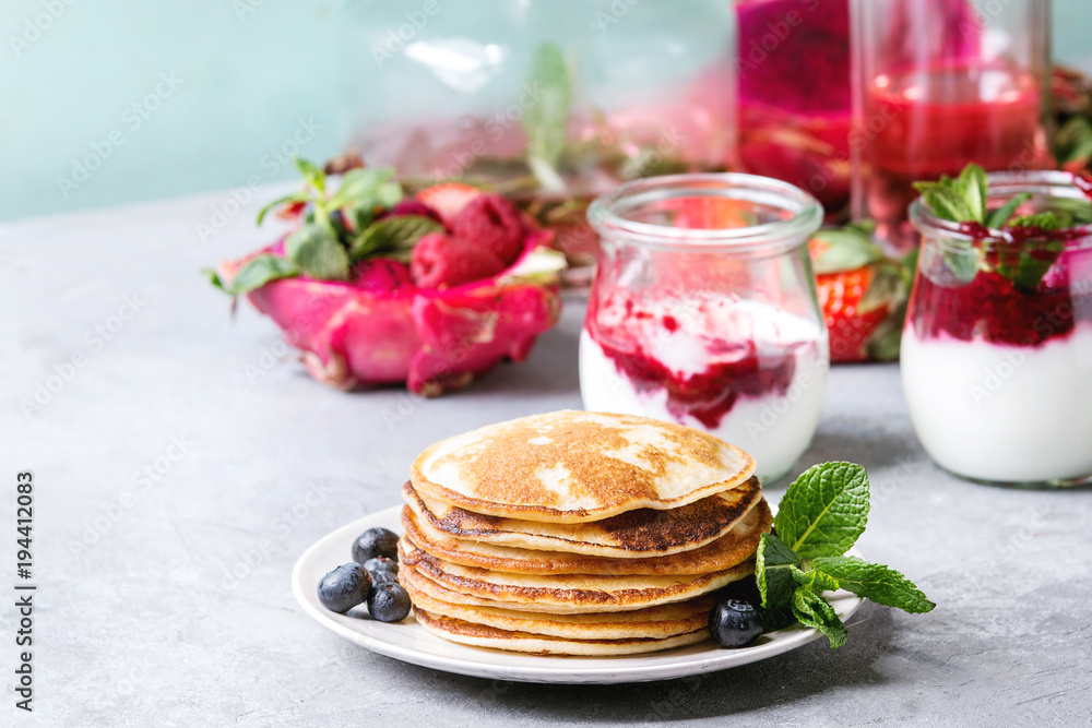 Stack of homemade pancakes served on plate with berries, mint, glass jars of yogurt, bottle of lemonade, fruit salad in pink dragon fruit over grey texture table.