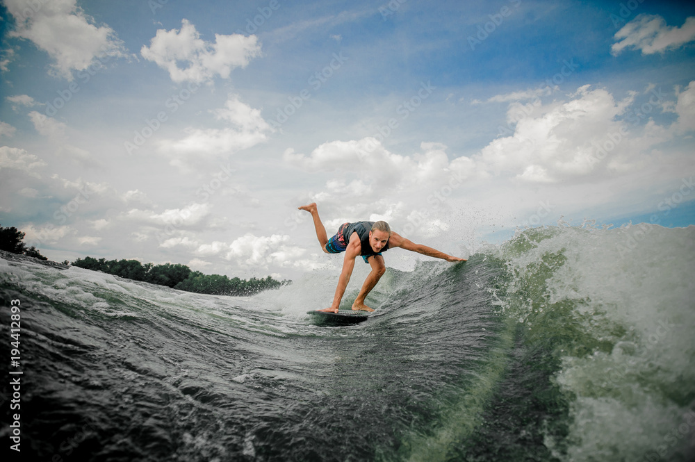 Young and athletic man wakesurfing on the board down the river against the sky