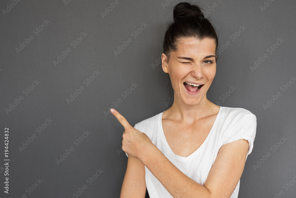 Portrait of young brunette Caucasian female wears casual, showing something interesting at copy space gray wall for your information or advertising content, winking at the camera, pointing sideways Portrait of young brunette Caucasian female wears casual, showing something interesting at copy space gray wall for your information or advertising content, winking at the camera, pointing sideways
