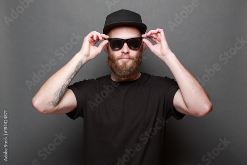 Hipster handsome male model with beard wearing black blank t-shirt with space for your logo or design over gray background