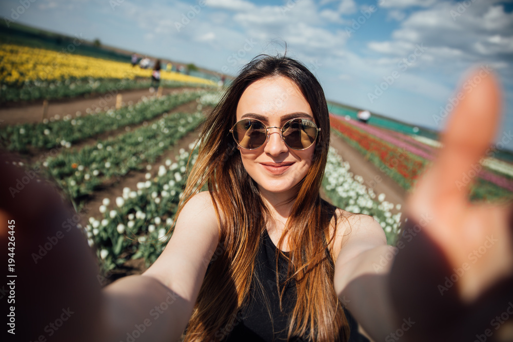 Fototapeta premium Young beautiful woman wear sunglasses make selfie in tulips flower field in sunny spring day