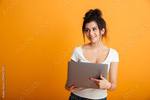 Photo of charming woman with long brown hair in bun holding silver personal computer and looking sideways, over yellow wall copy space