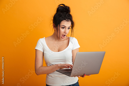 Photo of beautiful woman with long brown hair in bun holding and looking on silver laptop with surprise, over yellow wall copy space