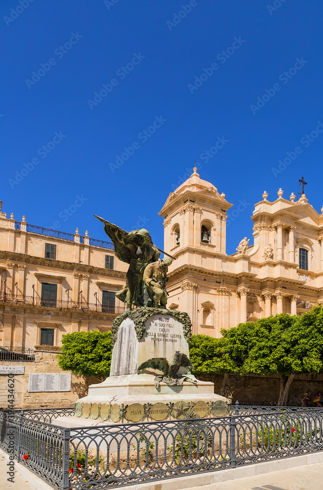 Noto, Sicily, Italy. Monument to the inhabitants of Noto, who died during the First World War, against the backdrop of the Palazzo Landolina and the Cathedral