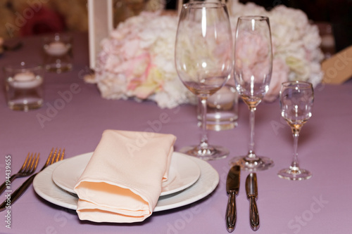 A stylish wedding decor of dishes, white bouquets with soft flowers, candles and glasses stand on a light tablecloth of a served table in a restaurant. Side view, selective focus