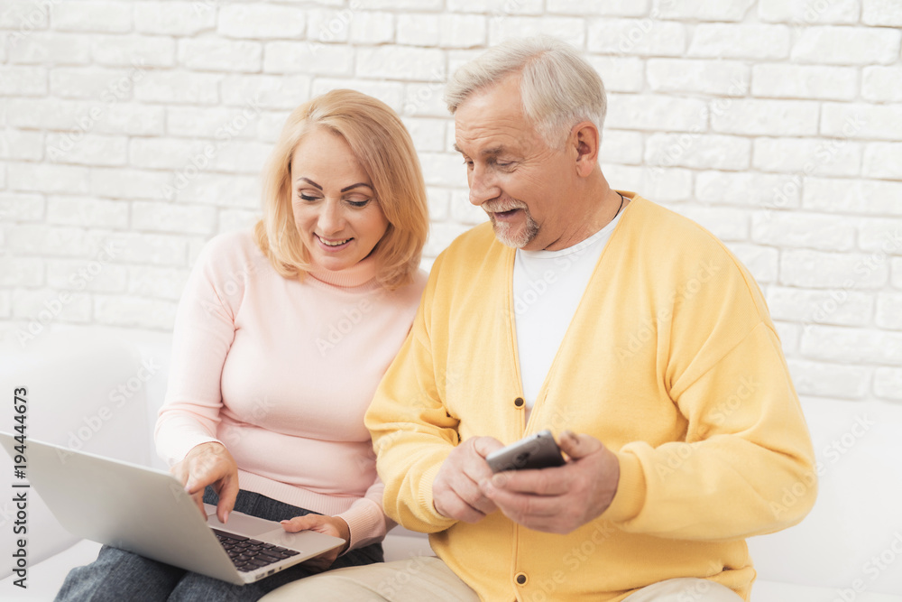 An older man and woman are sitting on a big white sofa and watching something on the laptop screen.