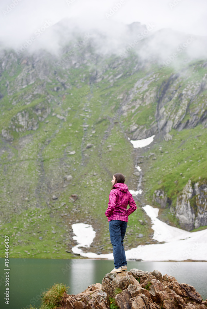 Pretty young girl hiker with hands in pockets standing on the edge of rocky cliff looking at the blue lake in the mountains in Romania. White clouds resting on mountain peaks, snow reflecting in water
