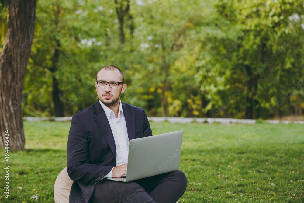 Young successful businessman in white shirt, classic suit, glasses. Man sit on soft pouf, working on laptop pc computer in city park on green lawn outdoors on nature. Mobile Office, business concept. Young successful businessman in white shirt, classic suit, glasses. Man sit on soft pouf, working on laptop pc computer in city park on green lawn outdoors on nature. Mobile Office, business concept.