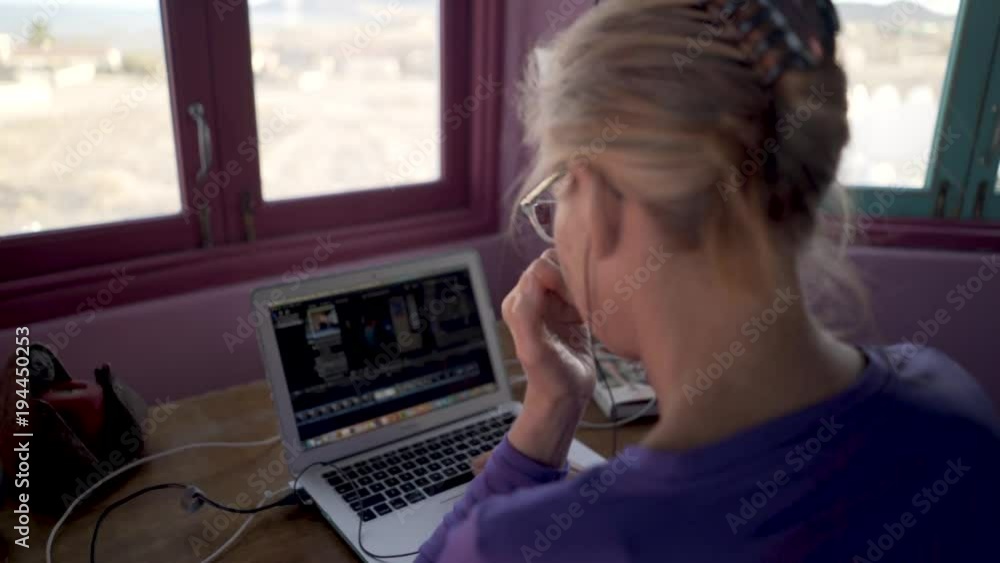 With earbuds and beautiful view a blond woman works on a movie on her laptop computer on a desk. With earbuds and beautiful view a blond woman works on a movie on her laptop computer on a desk.