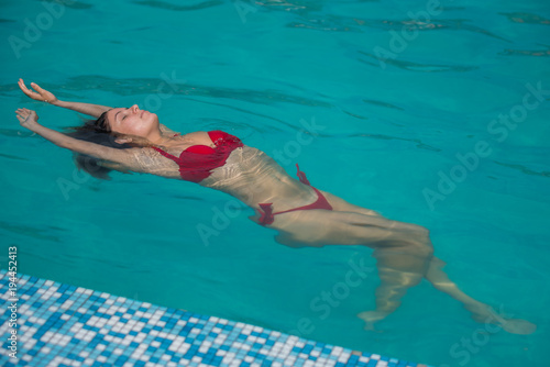 young attractive woman in red swimwear in pool with head thrown back side view. Young sexy woman with pretty face and wet hair swimming in pool with blue water sunny summer day outdoor