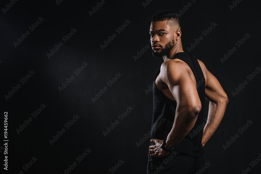 handsome young african american sportsman standing with hands on waist and looking at camera isolated on black