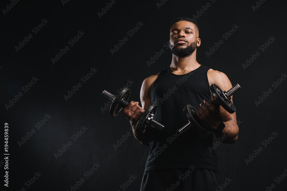 Fototapeta premium low angle view of young african american sportsman holding dumbbells and looking at camera isolated on black