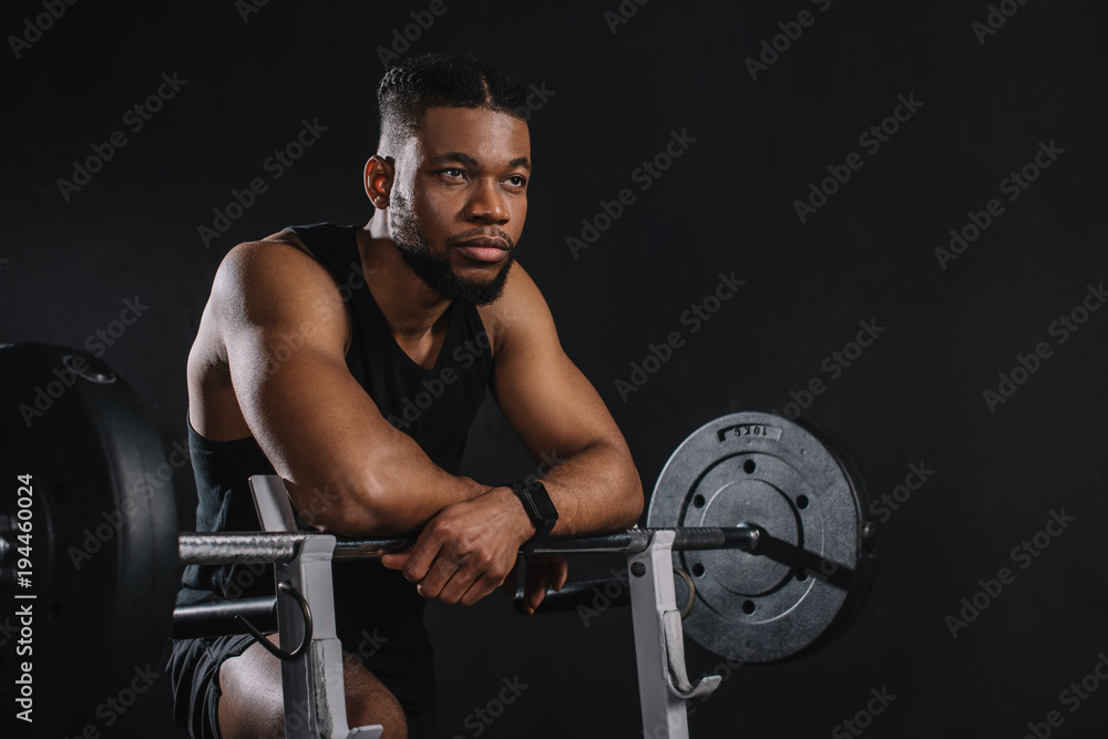 muscular young african american sportsman leaning at barbell and looking away on black muscular young african american sportsman leaning at barbell and looking away on black