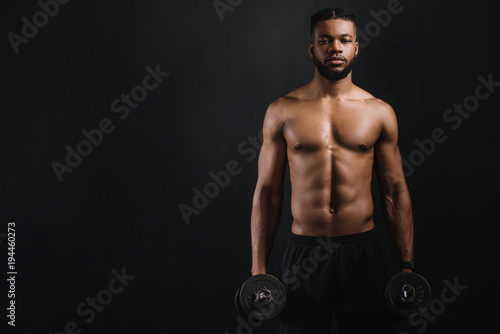 muscular shirtless african american man holding dumbbells and looking at camera isolated on black
