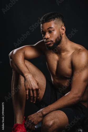 pensive shirtless young african american sportsman sitting and looking away on black