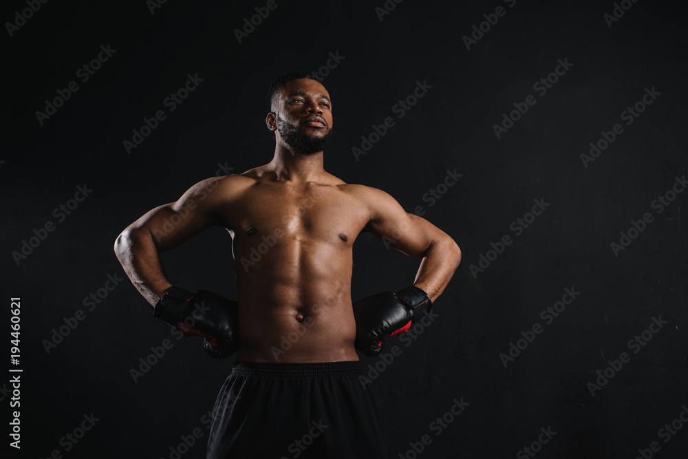 muscular shirtless african american man in boxing gloves standing with hands on waist and looking away isolated on black