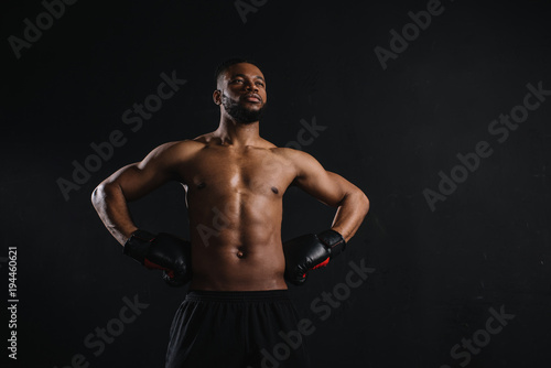 muscular shirtless african american man in boxing gloves standing with hands on waist and looking away isolated on black