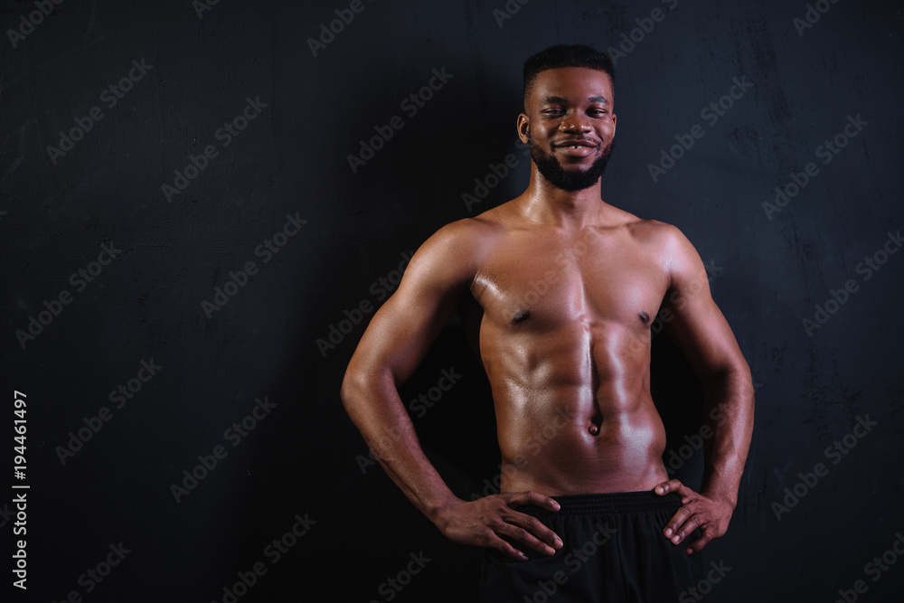 shirtless muscular young african american man standing with hands on waist and smiling at camera on black