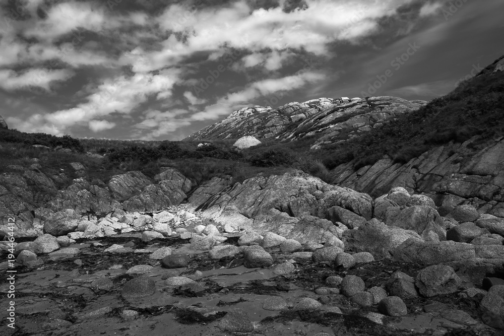 Ancient granite rocky landscape on the coast of east Scotland UK. Monochrome wide angle panorama - small rocks on a beach in the foreground rising up to the top of a distant hill.  