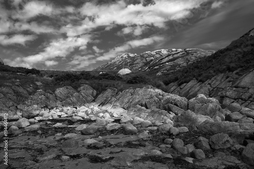 Ancient granite rocky landscape on the coast of east Scotland UK. Monochrome wide angle panorama - small rocks on a beach in the foreground rising up to the top of a distant hill. 