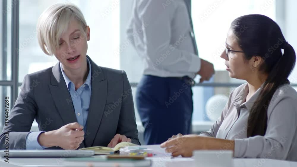 Beautiful businesswomen smiling and discussing notes in organizer at office desk; two businessmen talking in background Beautiful businesswomen smiling and discussing notes in organizer at office desk; two businessmen talking in background