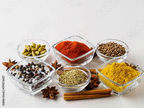spice set-coriander, red pepper, turmeric, cinnamon, star anise, rosemary various seasonings in glass cups, on white wooden table, side view, blank space for text.