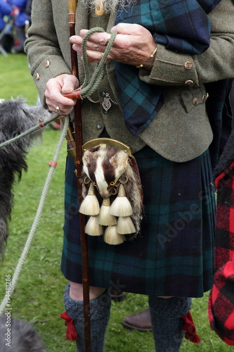 A Scottish man with an unusual sporran featuring a badgers head wearing a traditional kilt and tweed jacket . Highland clothing. Dark green and blue kilt green tweed jacket.