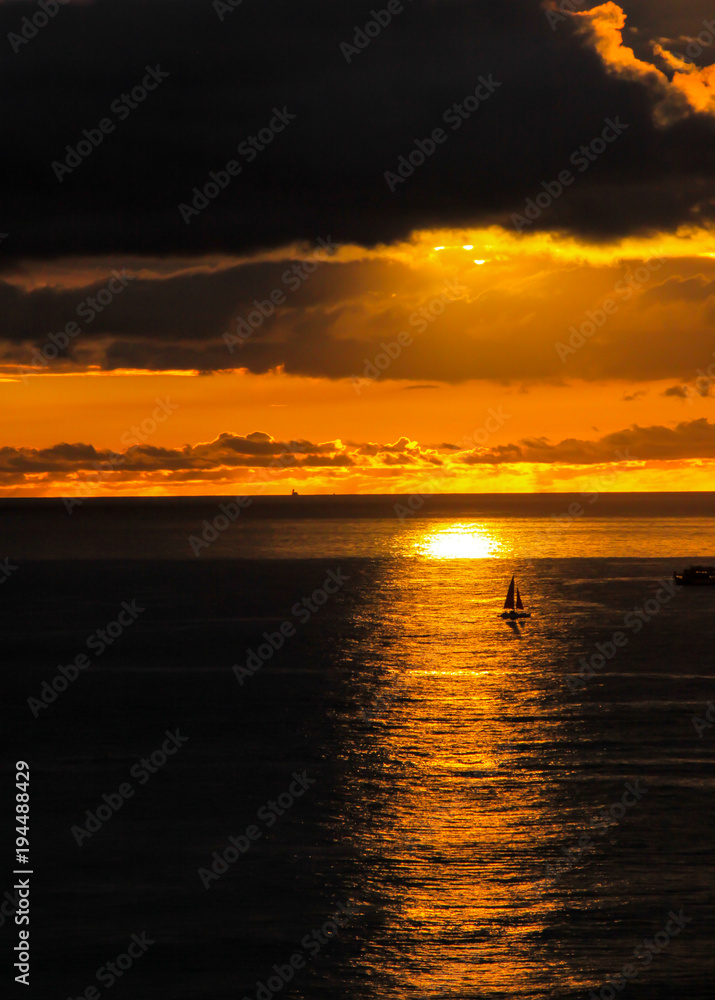 Sailboat silhouetted against the Sun's reflection under an orange sky.