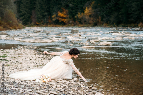 Beautiful dark-haired bride in beige dress with wedding bouquet on the riverside, mountains and pines on background