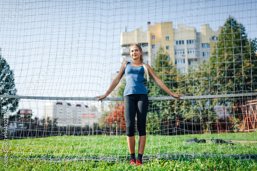 Beautiful stylish woman in a blue shirt and leggings stands near a football goal at the stadium at sunset. Beautiful sunlight. A girl with a perfect figure and in great shape. © lena_itzy