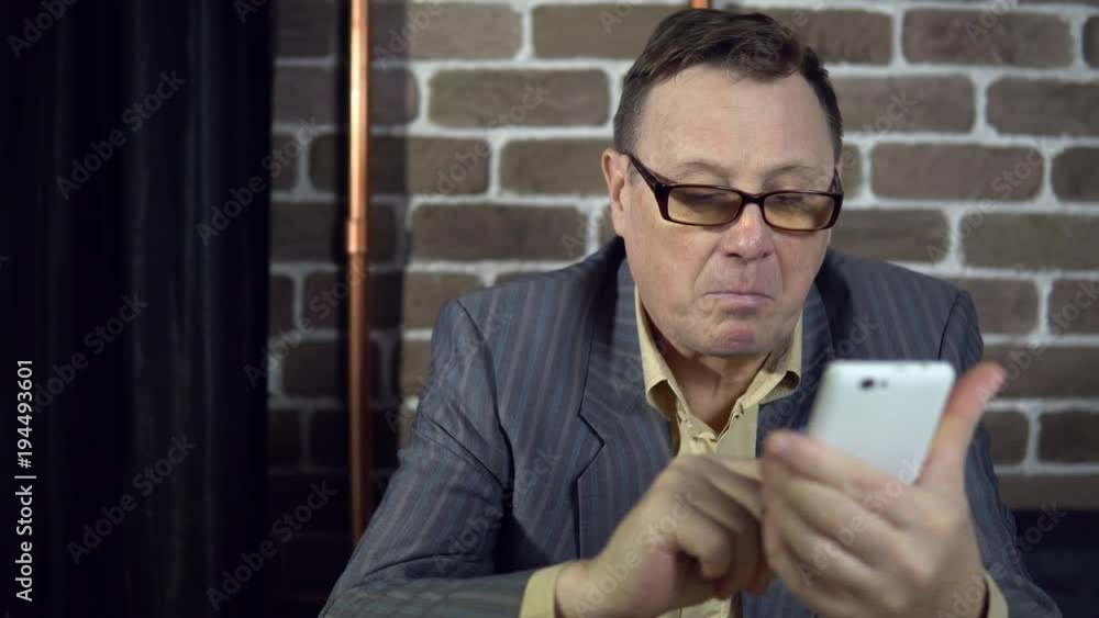 A busy elderly businessman in glasses uses a white smartphone, typing messages, reads articles, is surprised at the news.