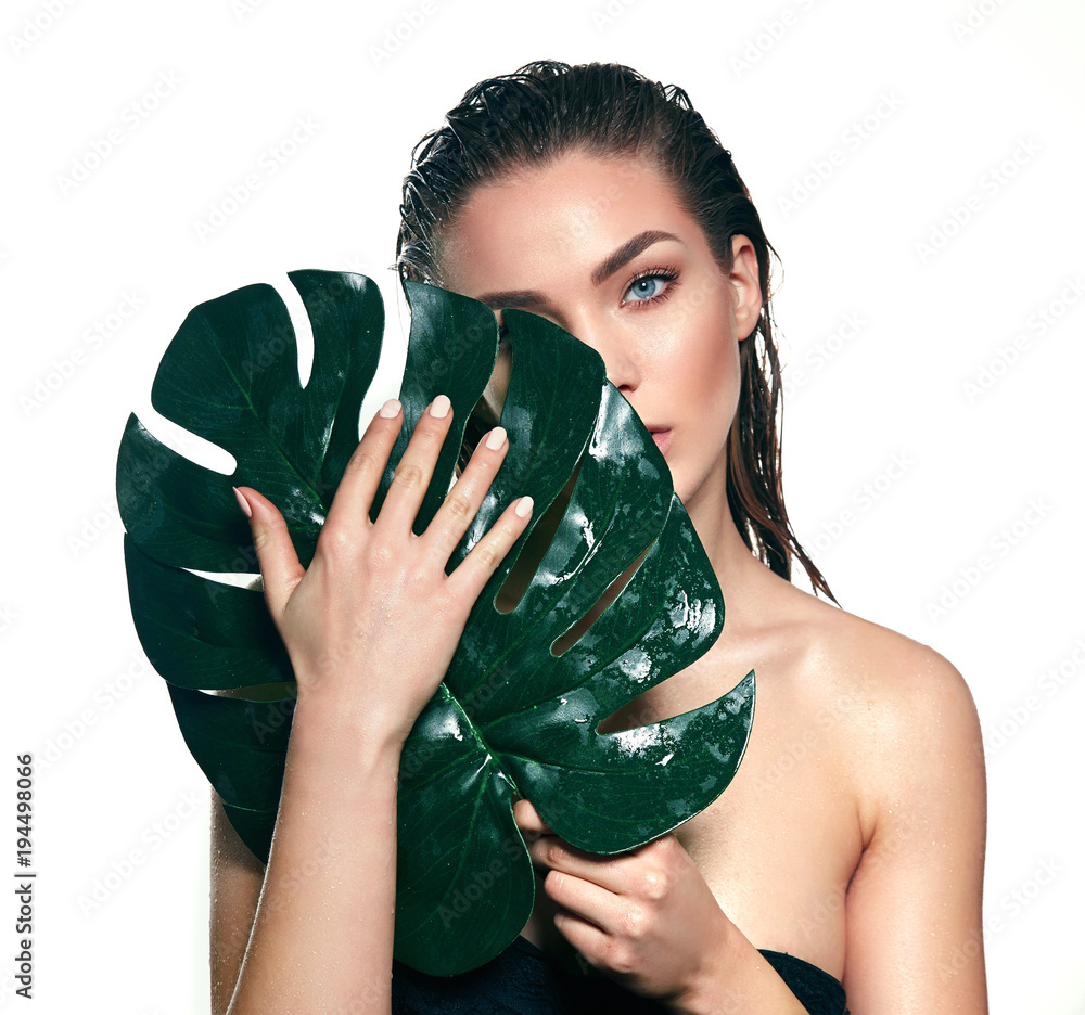 Obraz premium A beautiful young girl in the studio on a white background with wet skin and wet hair holds a large green tropical leaf in hands and covers a part of her face.