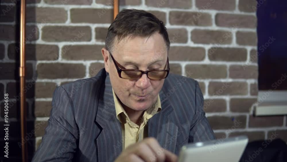Senior businessman in eyeglasses is working with a digital tablet at the office with a brick wall. Senior businessman in eyeglasses is working with a digital tablet at the office with a brick wall.