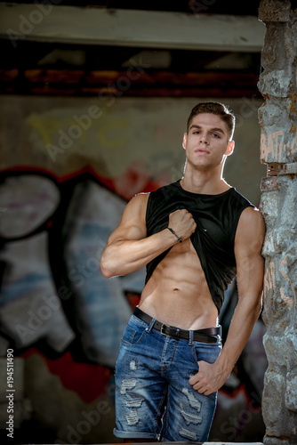 Muscled young man lean on rock wall lifting shirt and show his abs , hand in the pocket