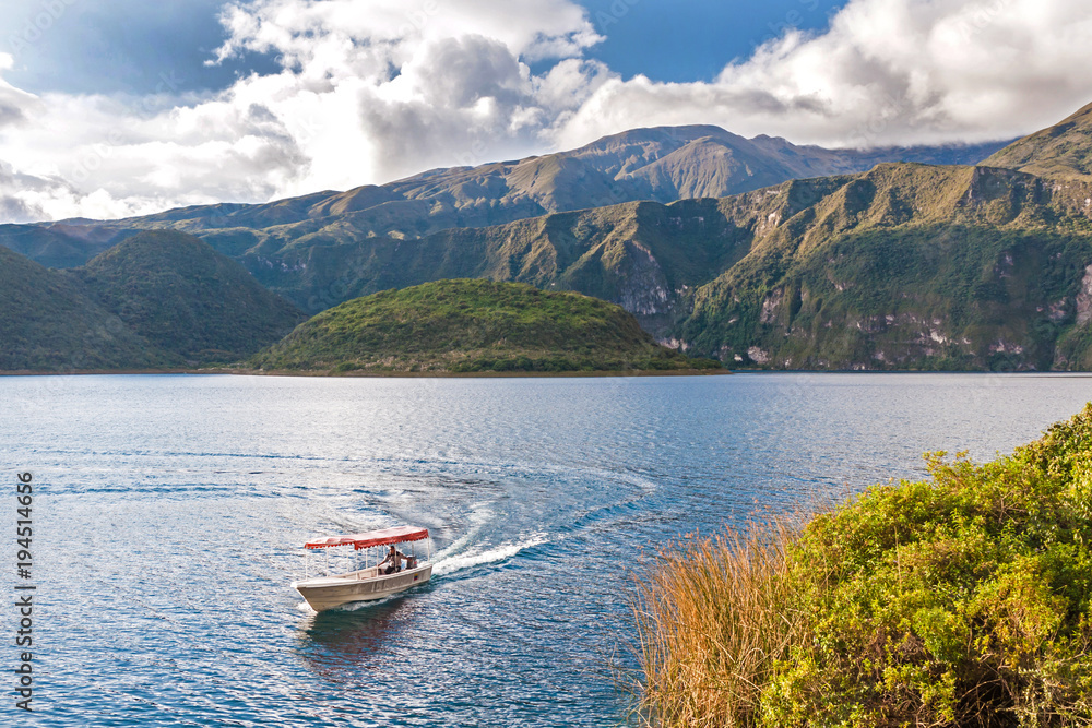 View of the Cuicocha lake and crater, with a small touristic boat in the water, on a sunny and cloudy afternoon.